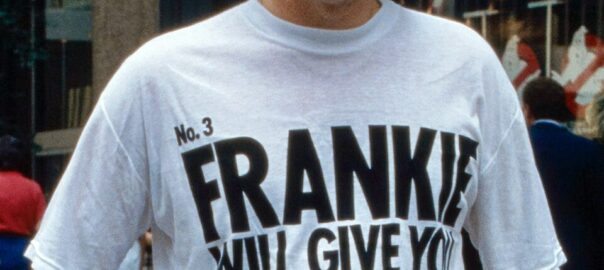 Trevor Horn en 1984 dans une rue de New York, avec des lunettes et un t-shirt blanc au slogan de Frankie Goes to Hollywood